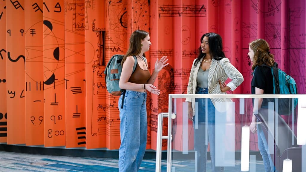 Three students stand near the stairs in D.H. Hill Jr. Library with a wall behind them painted in a rainbow gradient.