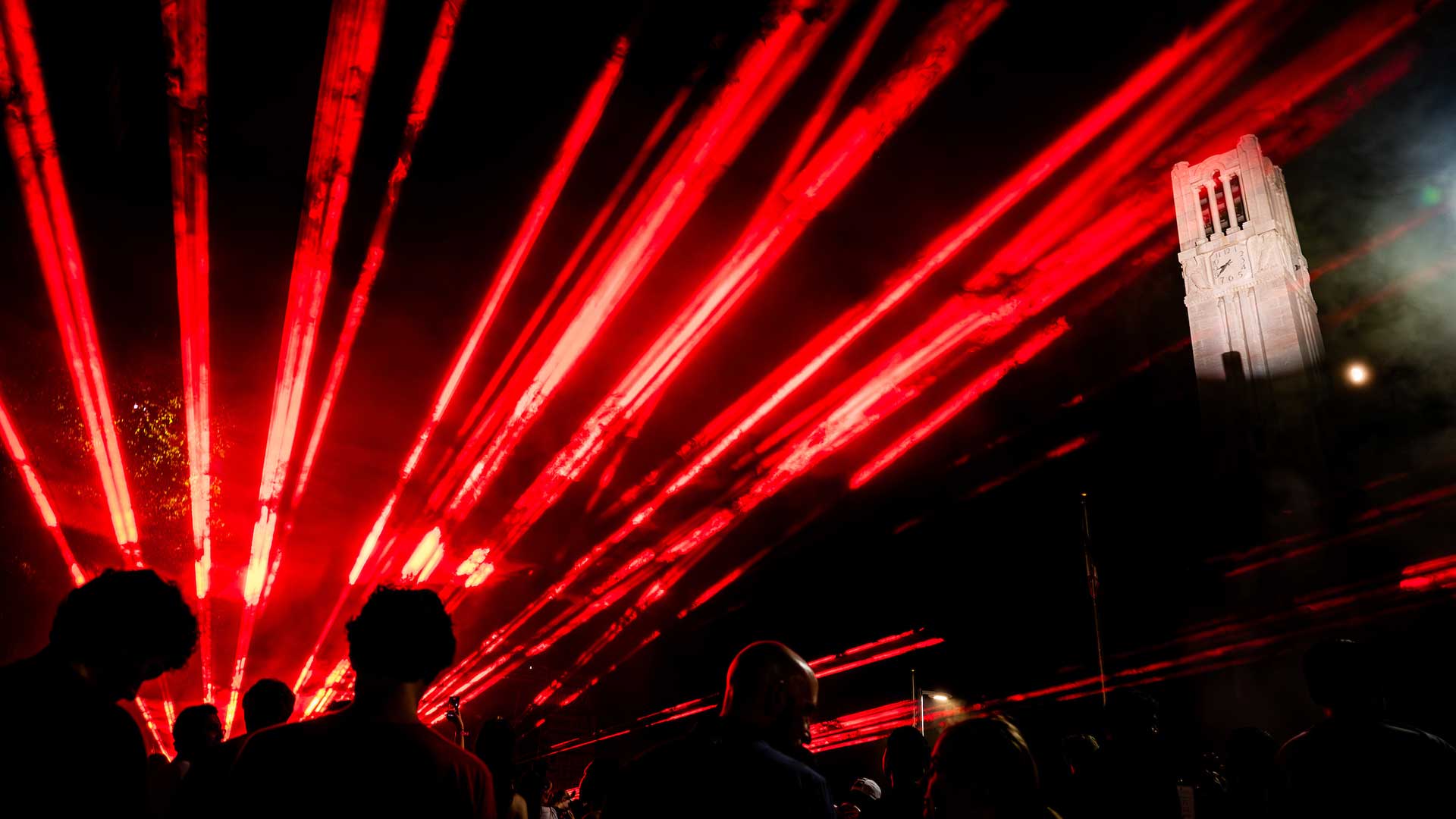 Red laser lights stream through the night sky above a crowd of people, with the Memorial Belltower illuminated in the background.