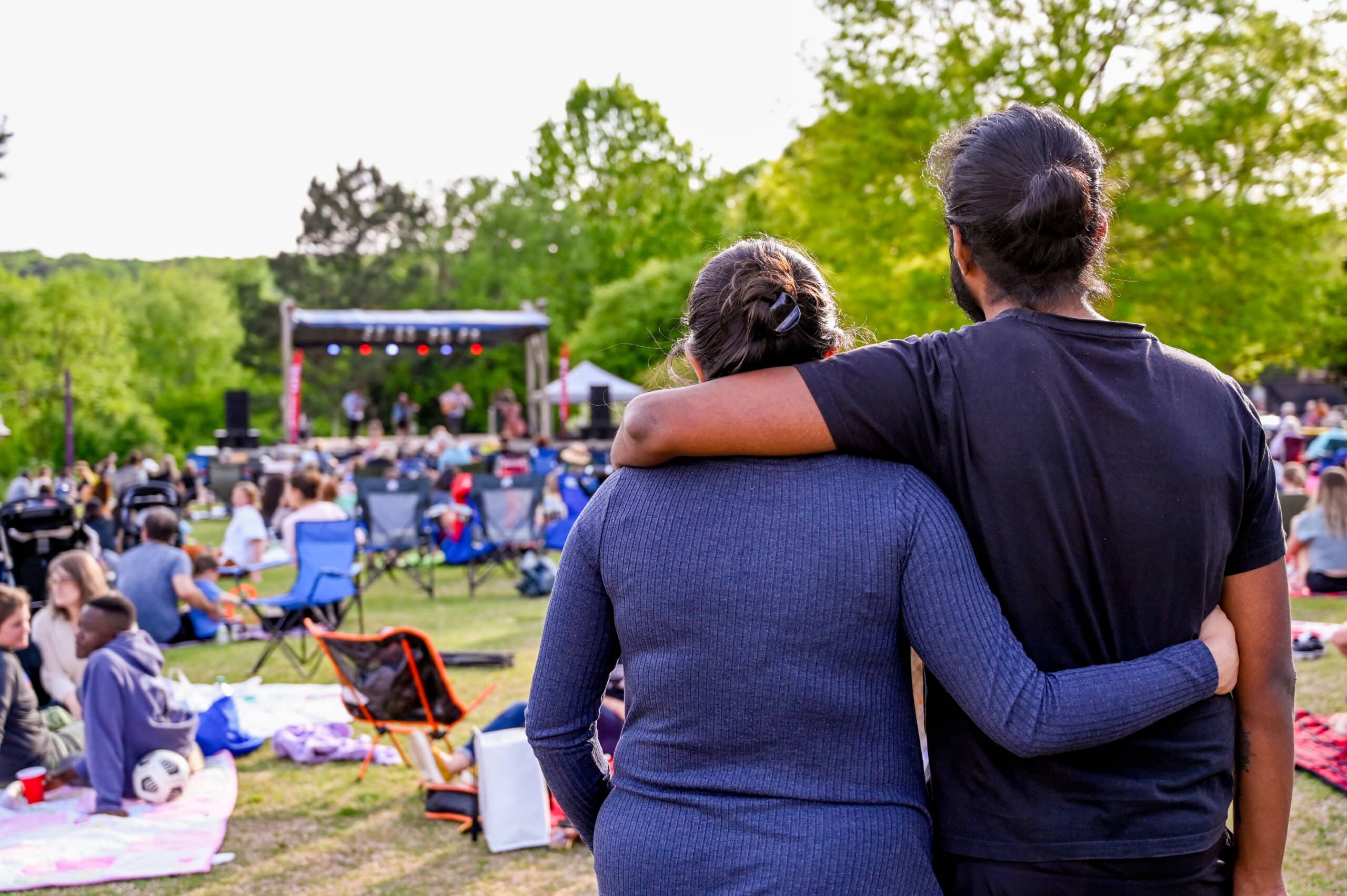 A crowd sits on the lawn during a Live@Lake Raleigh event on Centennial Campus, with two people standing together in the forefront of the image.