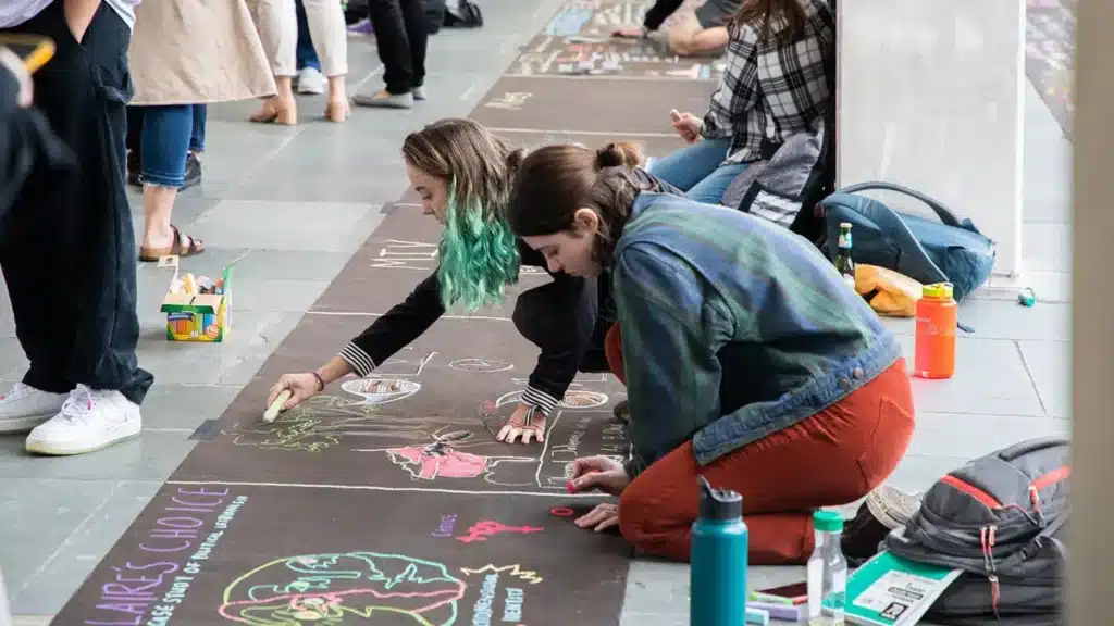 Two students draw on the sidewalk using colorful chalk.