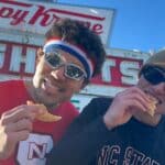 Two students hold half-eaten donuts in front of the Krispy Kreme sign.