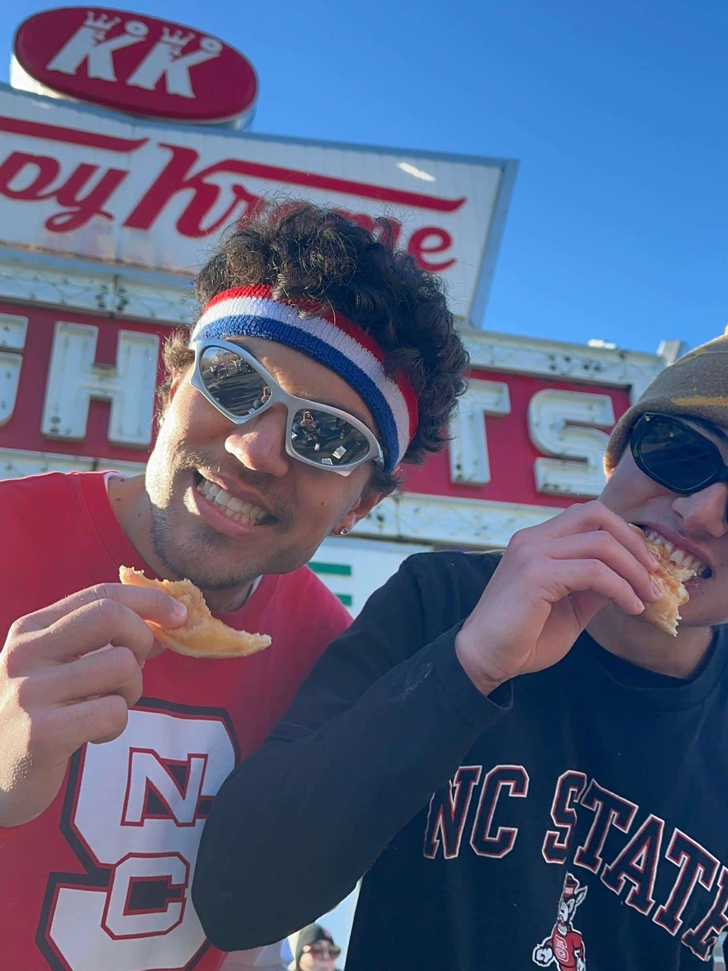 Two students hold half-eaten donuts in front of the Krispy Kreme sign.